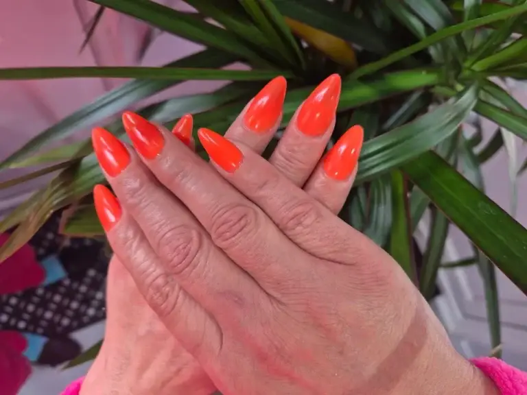 Bright orange pointed manicure with green plant background at Bhawani Beauty Guildford